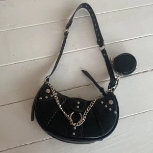 Svart axelväska med kedja från Soya - Cool svart axelväska från Soya Fashion i syntetmaterial. Halvmåneformad modell med silverfärgade nitar, kedjedetalj och dragkedja upptill. Väskan har justerbart axelband och en liten rund miniväska på sidan. Perfekt för att lyfta din outfit.