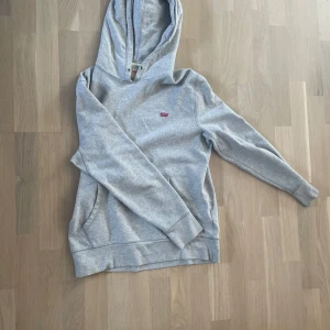 Grå hoodie från Levi's med huva - Snygg grå hoodie från Levi's i storlek S. Klassisk modell med huva, magficka och diskret Levi's-logga på bröstet. Tillverkad i mjuk bomullsmix som är perfekt för chill dagar. Långärmad och med ribbade muddar för extra komfort.