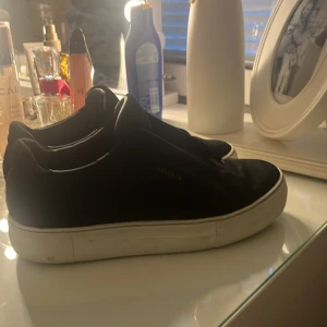 Svarta sneakers från Axel Arigato - Stilrena svarta sneakers från Axel Arigato med vit platt sula och diskret guldfärgad logga på sidan. Ovandelen är i mocka och skon har elastiskt band över vristen istället för klassisk snörning. Perfekta för en clean och modern look.