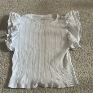 Vit ribbad topp med puffärm Gina Tricot - Supersöt vit topp från Gina Tricot med ribbat tyg och puffiga korta ärmar. Den har en rund halsringning och snygga vertikala ränder i strukturen. Perfekt för dig som gillar en stilren men ändå trendig look.