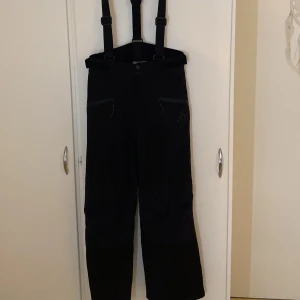 Svarta täckbyxor McKinley 152 - Svarta täckbyxor från McKinley med justerbara hängslen och dragkedjeförsedda fickor. Byxorna har en clean look med diskret logga på benet och är perfekta för vinterns alla äventyr. Materialet är slitstarkt och vattenavvisande syntet.