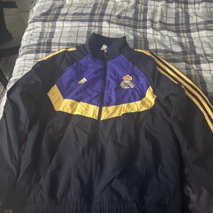 Real Madrid vindjacka från Adidas - Snygg Real Madrid vindjacka från Adidas i mörkblått med gula detaljer och klubbens emblem på bröstet. Jackan har dragkedja, klassiska Adidas-ränder på ärmarna och elastiska muddar. Perfekt för dig som vill visa ditt stöd för laget med stil.