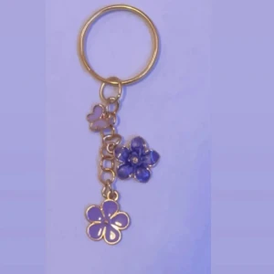 Guldigt blommigt nyckelring med hängen - Superfint nyckelring med guldfärgad kedja och tre blommiga hängen i lila och blått. Perfekt accessoar för att ge din väska eller nyckel en söt och lekfull touch. Blommorna har olika former och storlekar för en unik look.