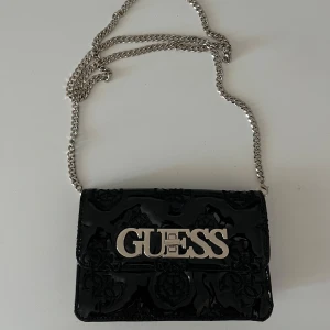 Svart axelväska från Guess med kedja - Snygg svart axelväska från Guess i glansigt skinnliknande material med en stor metallogo framtill. Väskan har silverfärgad kedjerem och praktiskt fack inuti. Perfekt för att lyfta din outfit med en trendig touch. HELT oanvänd och äkta!