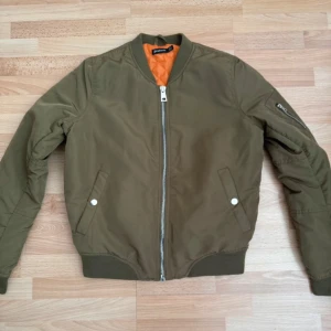 Olivgrön bomberjacka från Stradivarius - Säljer en olivgrön bomberjacka från Stradivarius med orange quiltat foder. Jackan har klassisk passform, dragkedja framtill, fickor med tryckknapp och en extra ficka med dragkedja på ärmen. Perfekt för dig som gillar streetstyle och vill ha en snygg look. Använd 2 gånger.