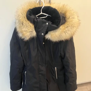 Svart vinterparkas med fuskpäls från Zara - Säljer en svart vinterparkas från Zara med stor fluffig fuskpäls runt huvan. Jackan har dragkedja, knappar och två fickor med dragkedja. Insidan är fodrad med mjuk svart fuskpäls och den har snörning i midjan för snygg passform. Perfekt för kalla vinterdagar! Nytt pris 1500kr.