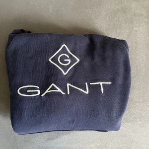 Mörkblå hoodie från GANT - Snygg mörkblå hoodie från GANT med stor vit logga på bröstet. Tröjan har huva, magficka och långa ärmar med ribbade muddar. Tillverkad i mjuk bomull som är skön att ha på sig. Perfekt för chill eller häng med kompisar.