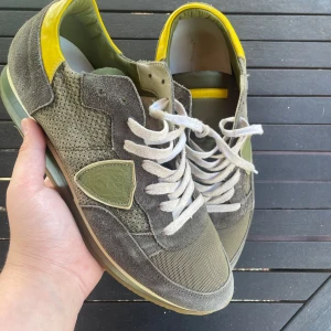 Gröna philipe models! - Snygga sneakers från Philippe Model i olivgrönt mocka och mesh, med vita snören och coola gula detaljer på häl och plös. Skorna har en klassisk låg siluett och en platt sula, samt märkets ikoniska sköld på sidan. Perfekta för dig som gillar streetstyle med en twist.