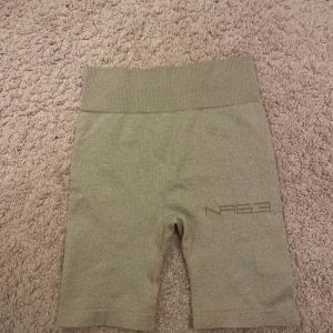 Beige ribbade cykelshorts NA - Snygga beige cykelshorts från New Apparel i ribbat material. De är högmidjade och har en tight passform som sitter skönt. Perfekta för träning eller när du vill ha en sportig look. Diskret logga på ena benet och stretchigt tyg för extra komfort.