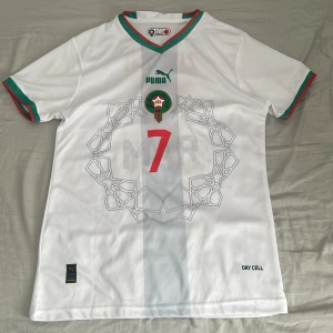 Marocko Ziyech #7 Puma fotbollströja - Snygg vit Marocko fotbollströja från Puma med Ziyech och nummer 7 på ryggen. Tröjan har gröna och röda detaljer vid krage och ärmslut, samt ett grafiskt mönster framtill. Tillverkad i lätt och ventilerande polyester med Dry Cell-teknologi.
