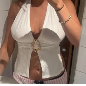 Vit halterneck topp med gulddetalj - Vit halterneck topp med djup v-ringning och en stor, vågig guldfärgad ring framtill. Toppen knyts i nacken och ryggen för perfekt passform. 🤍