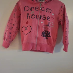 Rosa hoodie med Mumin-tryck och hjärtan - Söt rosa hoodie från Cantarana med dragkedja, huva och tryck av Lilla My samt texten 'Dream house'. Jackan har hjärtan och små muminmotiv över hela tyget. Perfekt för dig som gillar gulliga detaljer och vill ha en mysig och lekfull stil. Nopprig på några ställen. 
