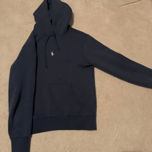 Mörkblå hoodie från Polo Ralph Lauren - Snygg mörkblå hoodie från Polo Ralph Lauren med klassisk vit broderad logga på bröstet. Tröjan har huva med snörning och en stor magficka. Perfekt för dig som gillar stilren och enkel streetwear.
