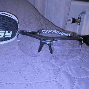 Innebandy glasögon  - Säljer ett par Unihoc sportglasögon med svart front och transparenta linser. Skalmarna har camouflage-mönster i grönt, beige och brunt samt justerbart band för extra säkerhet. Perfekta för innebandy eller annan sport. Fodral medföljer.
