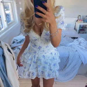 Blommig vit kortklänning med volanger - Supersöt vit kortklänning med blått blommönster, v-ringning och puffärmar. Klänningen har flera lager volanger i kjolen och en figurnära passform upptill. Köpt på plick förra året! Skriv för mer frågor och funderingar 💕