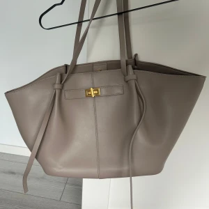 Beige axelväska med gulddetalj från Gina  - Stilren och rymlig beige axelväska i slätt skinn med långa handtag och snygg guldfärgad vridknäppning framtill. Väskan har minimalistisk design och passar perfekt för dig som vill ha en clean och modern look. Endast andvänd en gång 