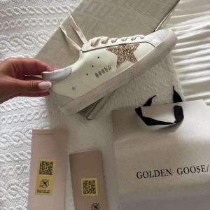 Golden Goose sneakers med glitterstjärna - Snygga sneakers från Golden Goose i vitt skinn med beige mockadetaljer och en glittrig stjärna i guld på sidan. Skorna har rund tå, platt sula och klassisk snörning. Perfekta för dig som vill sticka ut med en cool detalj.