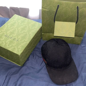 Svart keps från Gucci  - KVITTO FINS. Snygg svart keps från Gucci med klassiskt GG-monogram över hela kepsen och gröna/röda detaljer på sidan. Perfekt accessoar för att lyfta din outfit med exklusiv känsla. Kepsen har böjd skärm och justerbar passform. 