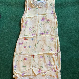 Blommig ärmlös klänning KappAhl  - Superfin ärmlös klänning från KappAhl i ljusgul ton med blommigt mönster i lila, rött och grönt. Luftigt material, perfekt för varma dagar. Rak passform och rund halsringning. Snygg till sneakers eller sandaler!