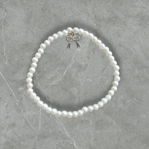 Pärlarmband med rosettberlock - Sött armband med vita pärlor och en liten silvrig rosettberlock. Perfekt accessoar för att ge din outfit en gullig och elegant touch. Pärlorna är jämna och sitter på en elastisk tråd som gör det enkelt att ta på och av.