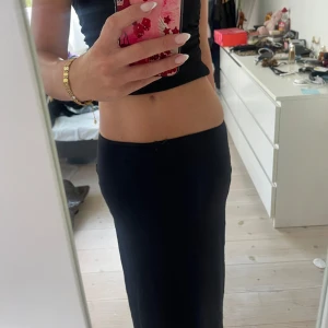 Svart långkjol med slits - Säljer en svart långkjol med låg midja men går att använda den på olika sätt beroende på hur man vill att den ska sitta. Det är en  snygg slits på sidan. Kjolen har en stilren och enkel design som sitter tight över höften och faller rakt ner. Perfekt för dig som gillar minimalistisk stil och vill ha något som känns både coolt och bekvämt.