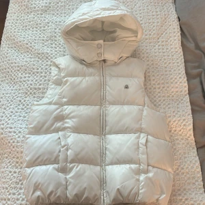 Vit dunväst med huva från Benetton - Snygg vit dunväst från Benetton med stor vadderad huva och två fickor framtill. Västen har dragkedja och två knappar vid halsen för extra skydd mot kylan. Perfekt att slänga över en hoodie för en cool look. Den har inga defekter och är helt ren. 