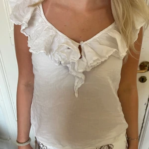 Vit volangtop med v-ringning - Vit linnetopp med volangdetaljer längs v-ringningen och axlarna. Toppen är ärmlös och har en liten knäppning framtill. 