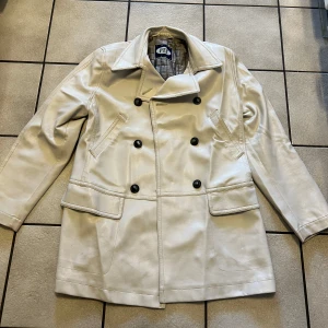 Beige skepparkavaj/jacka från PTA storlek M/L - Stilren beige skepparkavaj från PTA i skinnimitation med dubbelknäppning och stora svarta knappar. Jackan har klassisk krage, två snedställda fickor och två fickor med lock framtill. Perfekt för dig som vill ha en clean och tidlös look.