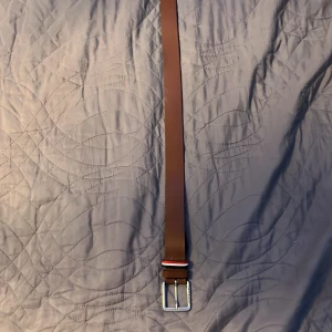 Brunt skärp från Jack & Jones - Snyggt brunt bälte från Jack & Jones i skinn med klassiskt silverfärgat spänne. Detalj med röd, vit och svart randig tygögla vid spännet ger en cool touch. Perfekt för att lyfta din outfit med en stilren accessoar.