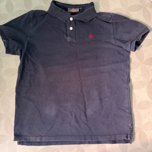 Mörkblå pikétröja från Morris - Klassisk mörkblå pikétröja från Morris med korta ärmar och krage. Tröjan har tre vita knappar framtill och en liten röd broderad detalj på bröstet. Perfekt för en stilren och avslappnad look.