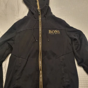 Svart hoodie från Hugo Boss - Svart hoodie från Hugo Boss med gulddetaljer. Jackan har dragkedja och huva med snörning. 