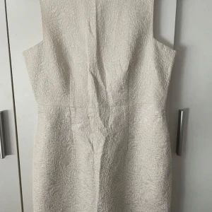 Elegant studentklänning  - Elegant beige klänning med strukturmönster och dragkedja i ryggen. Klänningen har en stilren design och är ärmlös, vilket gör den perfekt för olika tillfällen.