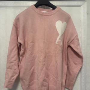 Rosa stickad tröja från Ami - Säljer en söt rosa stickad tröja från Ami med ett stort vitt hjärta på bröstet. Tröjan har en rund halsringning och långa ärmar. Perfekt för en mysig dag.