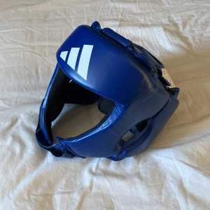 Boxningshjälm  Adidas - Säljer ett blått huvudskydd från Adidas, perfekt för kampsportsträning. Skyddet har en justerbar passform med kardborreband och är tillverkat i slitstarkt syntetmaterial. Den har aldrig använts alltså helt ny. 