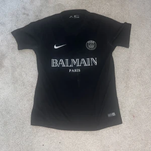 Svart PSG fotbollströja från Nike - Säljer en stilren svart PSG fotbollströja från Nike med Balmain Paris-logga på bröstet. Tröjan har korta ärmar och en rund halsringning. Perfekt för fotbollsfans som vill ha något unikt och trendigt. Passar både till match och träning.