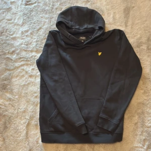 Svart hoodie från Lyle & Scott - Säljer en stilren svart hoodie från Lyle & Scott med deras ikoniska gula logga på bröstet. Tröjan har en klassisk känguruficka och en bekväm huva. Perfekt för en avslappnad look.