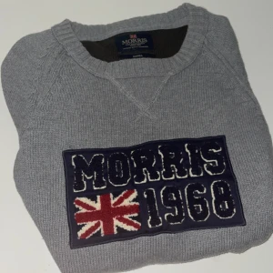 Grå tröja från Morris - Säljer en stilren grå tröja från Morris med ett broderat märke på bröstet som visar 'Morris 1968' och en brittisk flagga. Tröjan har en v-ringad hals och långa ärmar, perfekt för en avslappnad stil.