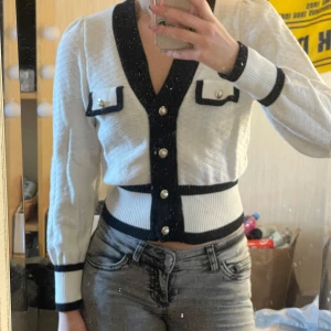 Vit och svart kofta med pärldetaljer - Säljer en stilren vit kofta med svarta detaljer och pärlknappar. Koftan har långa ärmar och dekorativa fickor framtill. Perfekt för en elegant look.