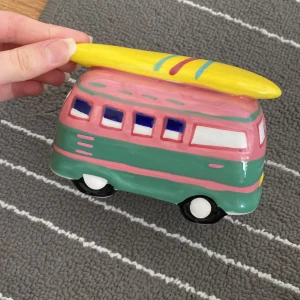 Keramisk sparbössa i form av en minibuss - Charmig keramisk sparbössa formad som en retro minibuss i rosa och grönt med en gul surfbräda på taket. Perfekt för att spara pengar med stil och en rolig inredningsdetalj för surfälskare. Designad i Danmark.
