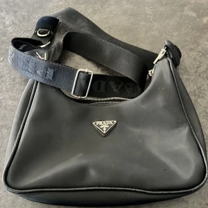 Svart axelväska från Prada - Snygg svart axelväska från Prada med justerbar axelrem och dragkedja. Väskan har en stilren design med Pradas logotyp på framsidan och ett mönstrat innerfoder. Perfekt för att bära dina dagliga nödvändigheter med stil. tyvärr gått sönder i ena knäppet till axelbandet men annars i helt nytt skick men säljer därför billigare