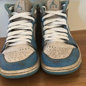 JORDAN 1 "UNIVERSITY BLUE GREY" MID - Snygga Nike Air Jordans i en fräsch kombination av blått och vitt. Skorna har klassisk snörning och en rund tå. Perfekta