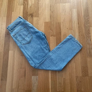 Blå jeans från Jack & Jones - Säljer ett par klassiska blå jeans från Jack & Jones. De har en tapered/regular fit och är tillverkade i denim med en snygg fransad kant vid benslutet. Perfekta för en avslappnad stil. Lite slitna längst ned på benen. 