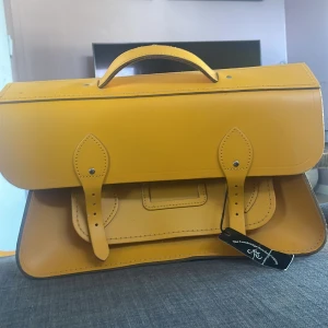 Gul skinnportfölj från The Cambridge Satchel Company - Säljer en stilren gul portfölj i högkvalitativt skinn från The Cambridge Satchel Company. Väskan har ett klassiskt utseende med ett handtag och spännen framtill. Perfekt för att bära dina viktiga dokument med stil.