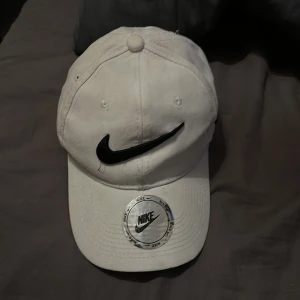 Vit keps från Nike - Snygg vit keps från Nike med den ikoniska svarta loggan framtill. Kepsen har en justerbar rem baktill för perfekt passform och är tillverkad i bomull. Perfekt för en sportig look eller en avslappnad dag ute. Köptes i 2021 för 200 eller 250kr och använd många gånger men är omhändertagen. Inuti är en autograf från Dogge Doggelito. 