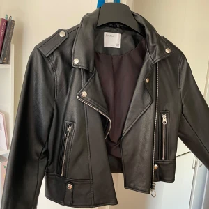 Svart skinnjacka från Bershka - Snygg svart skinnjacka från Bershka med dragkedja och knappar. Jackan har en klassisk bikerstil med axelklaffar och flera fickor med dragkedjor. Perfekt för en cool och edgy look. bar den bara tre gånger. Utan defekter. du betalar för leverans