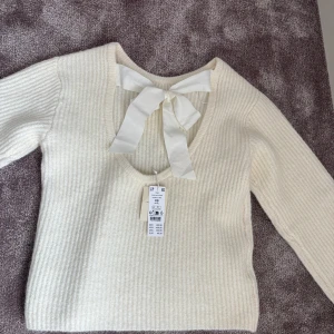 Vit stickad tröja med rosett - Säljer en söt vit stickad tröja med en stor rosett i nacken. Tröjan har en v-ringad design och långa ärmar. Perfekt för en stilren och mysig look. Passar bra till både jeans och kjol. Aldrig andvänd, prislapp är fortfarande på💞💞