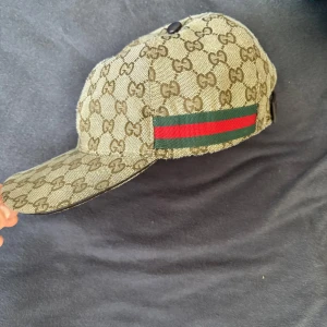Gucci keps - Snygg beige keps från Gucci med det ikoniska GG-mönstret. Kepsen har en grön och röd randdetalj på sidan och justerbar rem baktill. Perfekt för att ge din outfit en lyxig touch. Storlek: M