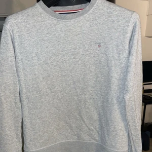 Grå sweatshirt från GANT - Säljer en stilren grå sweatshirt från GANT med broderad logga på bröstet. Tröjan har långa ärmar och en klassisk rund halsringning. Perfekt för en avslappnad look.