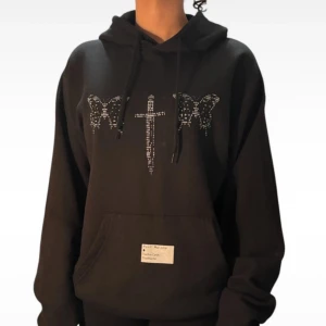 Svart hoodie med tryck från Post Malone - Snygg svart hoodie med ett unikt tryck av en dolk och fjärilar i glittrande detaljer. Hoodien har en klassisk passform med känguruficka och justerbar huva. Perfekt för dig som vill ha en stilren men ändå iögonfallande look. En merch från hans världsturné!