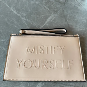 Beige kuvertväska från Mist - Säljer en stilren beige kuvertväska från Mist med texten 'MISTIFY YOURSELF' präglad på framsidan. Väskan har en praktisk handledsrem och stängs med dragkedja. Perfekt för att bära dina viktigaste saker med stil.